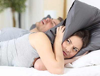 Frustrated woman covering her ears in bed with snoring man in need of sleep apnea therapy